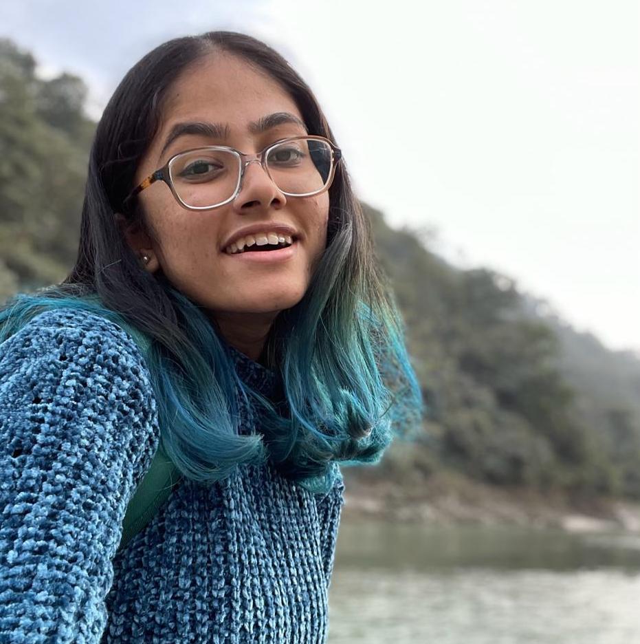 Mital smiling, wearing glasses. The bottom of her hair is dyed blue and she is in nature with a mountainous backdrop